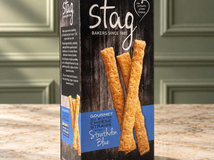 A box of Stag Bakers Strathbann Blue Gourmet Cheese Straws, featuring a dark wood-textured black box with blue and white text and images of golden-brown cheese straws, set on a granite countertop with a blurred kitchen background.